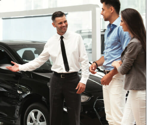 Man in formal attire showcasing cars on sales floor to a couple